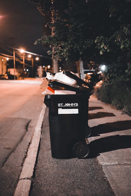 A black wheeled rubbish bin labeled 'ST. JOHN'S' is positioned on the edge of a paved sidewalk next to a street at night. The bin is open and filled with various waste items, including flattened cardboard boxes, plastic packaging, and paper debris, which overflow slightly over the top rim. The surface of the bin has a matte finish, with visible signs of dirt and usage. The street is illuminated by warm streetlights, casting elongated shadows of the bin and nearby trees onto the pavement. In the background, faint outlines of trees and residential buildings are visible, along with parked cars along the curb. The environment suggests a quiet urban or suburban area, where private waste disposal is occurring, distinct from municipal collection. The scene emphasizes the importance of proper rubbish handling and the role of independent waste collection services like those provided by rubbishremovalephantandcastle.org.uk in managing household waste outside of council services, especially during evening hours.