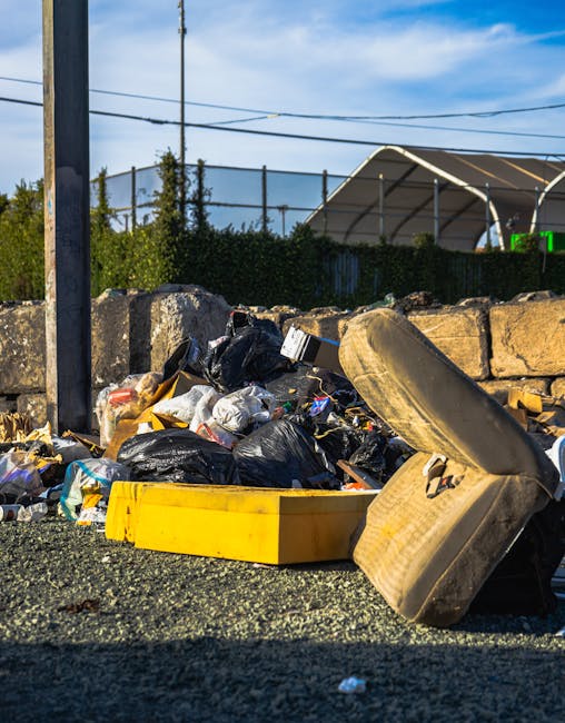 A pile of mixed household rubbish and waste materials are accumulated on a gravel surface next to a low stone wall. The rubbish includes several black plastic garbage bags, some partially torn open revealing trash inside, along with a yellow plastic storage container lying on its side. There is a large, weathered car seat with visible fabric texture and dirt, positioned leaning against the bags. In the background, behind the wall, environmental elements such as green hedges, a metal pole with electrical wires, and a half-visible structure with a curved roof can be seen under a bright blue sky with wispy clouds. The scene suggests a typical area for private waste disposal, possibly associated with independent rubbish removal services, rather than municipal collection. The overall environment is outdoors in an alley or driveway, indicating a non-public waste handling scenario. Occasionally, the scene emphasizes the importance of proper rubbish clearance to prevent improper or illegal dumping, aligning with the context of alternative waste removal options in the area.