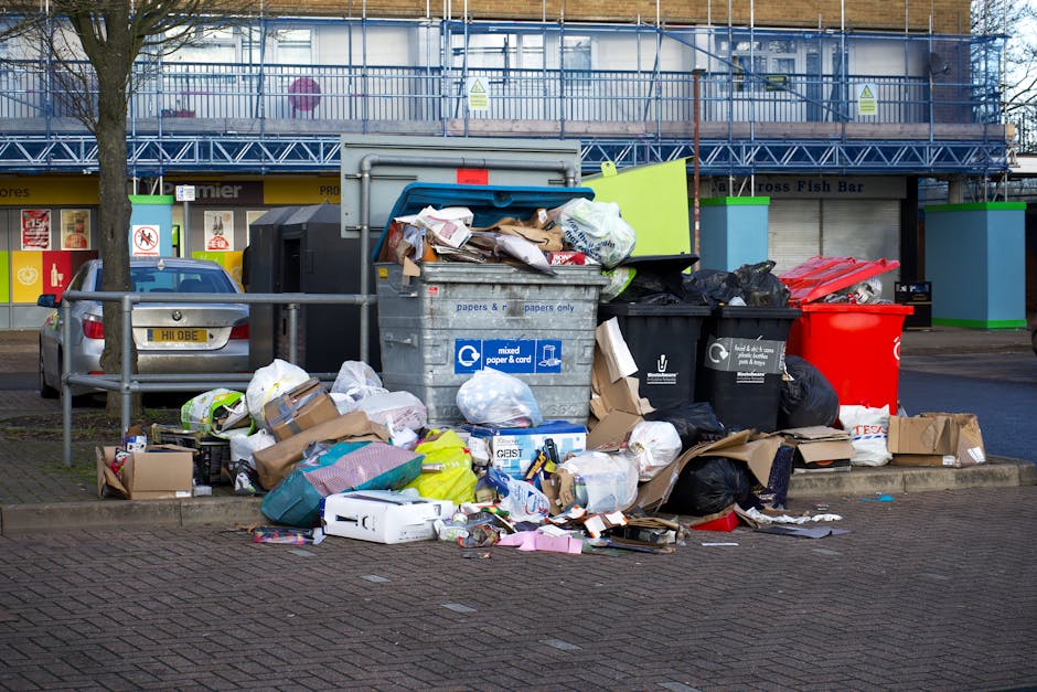 A black wheeled rubbish bin labeled 'ST. JOHN'S' is positioned on the edge of a paved sidewalk next to a street at night. The bin is open and filled with various waste items, including flattened cardboard boxes, plastic packaging, and paper debris, which overflow slightly over the top rim. The surface of the bin has a matte finish, with visible signs of dirt and usage. The street is illuminated by warm streetlights, casting elongated shadows of the bin and nearby trees onto the pavement. In the background, faint outlines of trees and residential buildings are visible, along with parked cars along the curb. The environment suggests a quiet urban or suburban area, where private waste disposal is occurring, distinct from municipal collection. The scene emphasizes the importance of proper rubbish handling and the role of independent waste collection services like those provided by rubbishremovalephantandcastle.org.uk in managing household waste outside of council services, especially during evening hours.