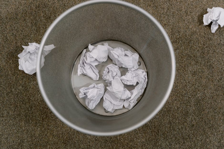 A cylindrical metal waste bin with a matte grey finish, positioned on a brown textured carpeted floor. Inside the bin are several crumpled sheets of white paper, some partially submerged in a small amount of water at the bottom. Outside the bin, on the carpet, there are additional crumpled paper pieces, indicating recent disposal. The environment suggests an indoor setting, possibly an office or workspace, where waste paper is collected for removal. The image highlights an easy-to-empty rubbish container used for paper waste, fitting within the scope of domestic or business rubbish collection services such as those offered by rubbishremovalelephantandcastle.org.uk, which could facilitate on-site or private waste handling for shops and offices.