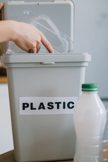 A close-up of a grey plastic recycling bin with a white label bearing the word 'PLASTIC' in bold black letters, situated on a wooden surface indoors. A hand is seen placing a clear plastic container into the open lid of the bin, which has a smooth, matte finish. To the right of the bin, there is a partially visible white plastic bottle with a green cap. The background features a light-colored wall, and the scene emphasizes the process of separating recyclable plastics as part of an environmentally-conscious rubbish removal practice within a domestic or office setting, highlighting the importance of proper waste sorting for effective rubbish management. The lighting is natural and evenly distributed, illuminating the objects clearly for visual identification and accessibility.