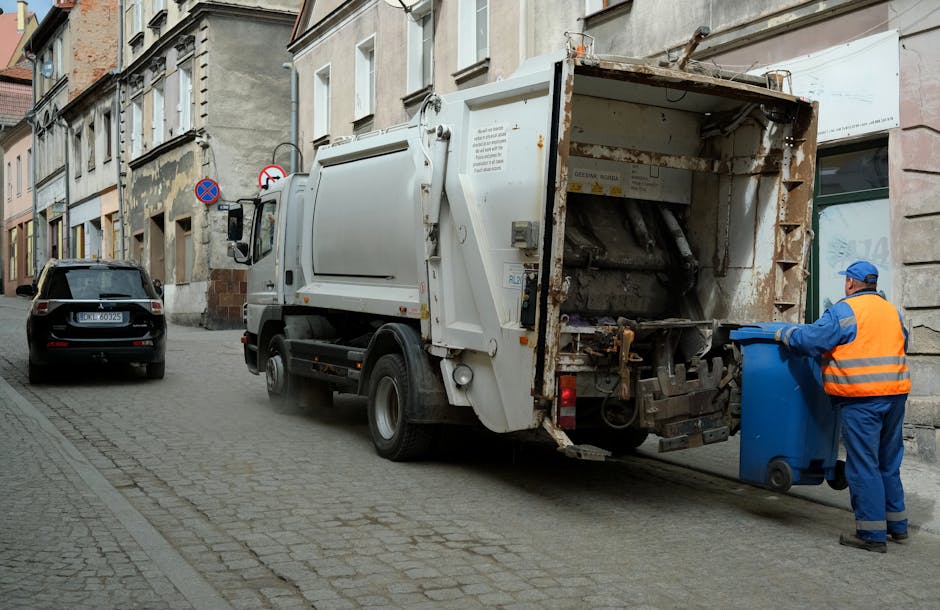 An outdoor scene showing a pile of mixed rubbish including black and transparent plastic bags, cardboard, paper, and a yellow plastic container, all scattered on a gravel surface. A portion of a worn, stained beige car seat or upholstered furniture is leaned against the debris. Behind the rubbish, there is a low stone wall made of irregularly shaped, rough-textured stones, with a green hedge and some plants visible beyond the wall. To the left, part of a metal pole and electrical wires overhead are visible, with a partly cloudy sky above. In the background, there is a large, curved, metallic or plastic structure, possibly a tent or sports facility roof, with a darkened interior and some green digital signage. The scene is lit by natural daylight, highlighting the textures and colors of the refuse and surroundings. The overall setting appears to be an informal rubbish collection or disposal site, indicative of alternative waste handling away from official waste services, aligning with independent rubbish removal practices.
