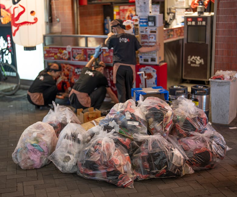 Several large transparent plastic bags filled with various types of waste, including clothing, paper, and miscellaneous debris, are stacked on a paved sidewalk at night. Behind the bags, three workers wearing dark uniforms and protective gloves are kneeling and sorting or collecting additional rubbish in front of a small fast-food outlet or shop. Some of the workers are leaning over boxes and containers, possibly organizing or handling waste materials. In the background, there are brightly lit signs, red-tiled walls, and equipment such as trash bins and recycling containers, indicating a commercial or densely populated area. The scene captures an organized effort in private waste handling or rubbish removal, with the focus on clearing accumulated refuse from the location. The overall lighting suggests evening hours, with artificial illumination highlighting the activity and surroundings, consistent with an independent rubbish collection process near a retail or dining establishment.