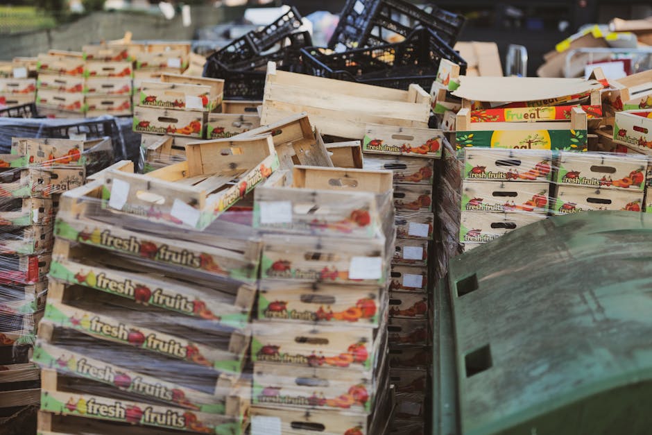 A large outdoor area filled with multiple stacks of empty wooden fruit crates, predominantly light-colored with printed labels displaying 'fresh fruits' and images of various produce. The crates are arranged in an irregular manner, some leaning, others stacked neatly, and a few overturned, revealing their open tops and side handles. Behind the crates, there are black plastic baskets and a pile of miscellaneous discarded objects, including cardboard boxes and packaging materials. To the right, a green, weathered plastic container, possibly a recycling bin or waste receptacle, is partially visible, with its surface showing signs of dirt and age. The scene appears to be situated on a paved surface, suggesting an outdoor marketplace or a dedicated waste collection area, with ambient lighting indicating daylight. This setting aligns with services related to independent rubbish collection or disposal of waste materials, such as those handled by rubbish removal specialists like rubbishremovalelephantandcastle.org.uk, emphasizing the physical nature of bulk waste or recyclable goods awaiting collection.