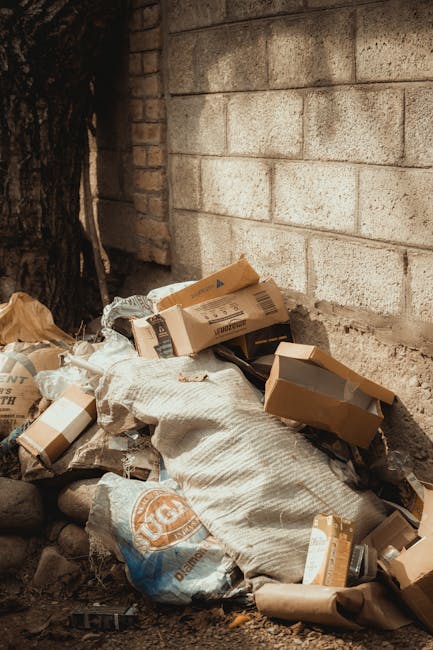 A pile of mixed rubbish, including flattened cardboard boxes, paper bags, and plastic packaging, is collected against a weathered brick wall and rough concrete surface. The cardboard boxes vary in size and appear partially crushed or torn, with some still bearing printed labels or barcodes. A large, textured white sack is partially buried under the debris, with its fabric showing signs of wear. Surrounding the rubbish are small stones and dirt on the ground, with a dark tree trunk visible on the far left side of the image. The scene is outdoors, with natural daylight illuminating the area, emphasizing the textures and colors of the waste materials. This images illustrates a typical example of waste accumulation that may be addressed through private rubbish removal services such as those offered by Rubbish Removal Elephant and Castle, highlighting the need for effective on-site clearance and disposal solutions within urban environments.