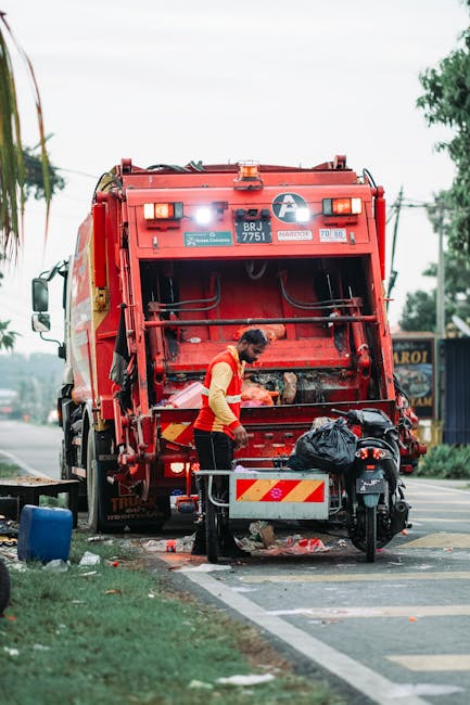 A worker dressed in a yellow safety vest with red detailing is engaged in rubbish collection at the rear of a large red waste disposal truck parked on the side of a street. The truck features a visible white circular logo and identification plates, with hydraulic mechanisms and a loading area that is open and empty awaiting waste. The worker is positioned next to a small collection of waste and tools placed on a metal cart, with a black garbage bag on top; the cart is located on the pavement near the vehicle. The scene is set outdoors under natural daylight, with overcast skies and surrounding elements including a blue container nearby, a parked motorcycle partially obscured by the specialist, and soft greenery and trees visible in the background. Power lines stretch across the sky, indicating an urban or semi-urban environment that supports independent waste collection activities, often used as an alternative to municipal rubbish removal services. The setting suggests a routine on-site rubbish clearance operation, aligning with private waste handling practices in a street environment.