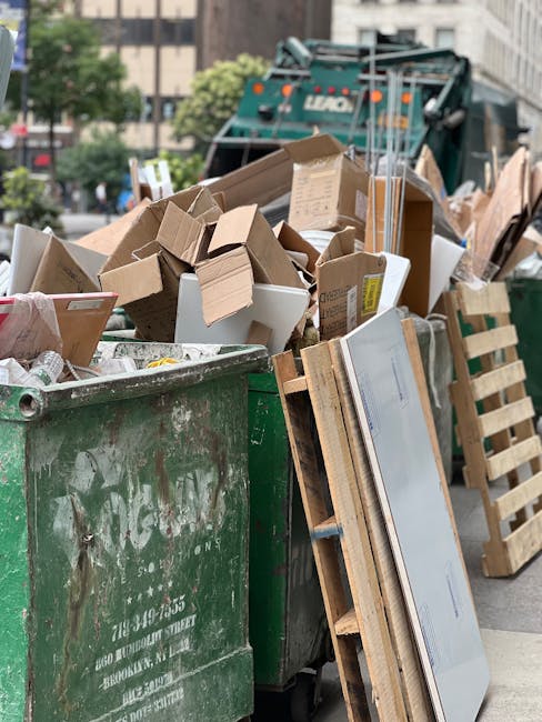 The image shows a large green municipal waste container filled with a mixture of cardboard boxes, some flattened and others still in box form, and various paper packaging. The cardboard exhibits different textures, from smooth and glossy to corrugated finishes, and the boxes are in shades of brown, with some having printed labels or warnings. Nearby, leaning against the container, are wooden pallets and a flat, panel-like object, possibly a piece of furniture or a large electronic device, with visible edges and a reflective surface. In the background, a commercial waste collection vehicle is partially visible, painted in dark green, with some text and logos on its side, suggesting a professional rubbish removal service. The scene is set outdoors on a street with a paved surface and urban buildings, with hints of trees and parked vehicles in the distant background. Natural daylight illuminates the scene, highlighting the textures and colors of the waste materials, which are indicative of typical household and small-scale commercial rubbish awaiting collection and disposal, aligning with independent or private waste handling services.